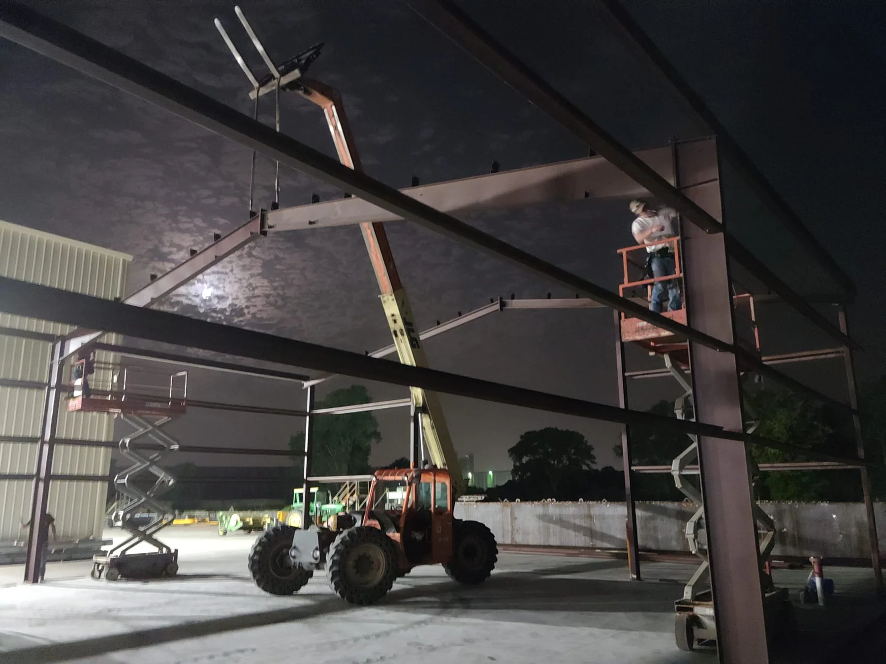 Workers on raised lifts beside steel framing during nighttime construction