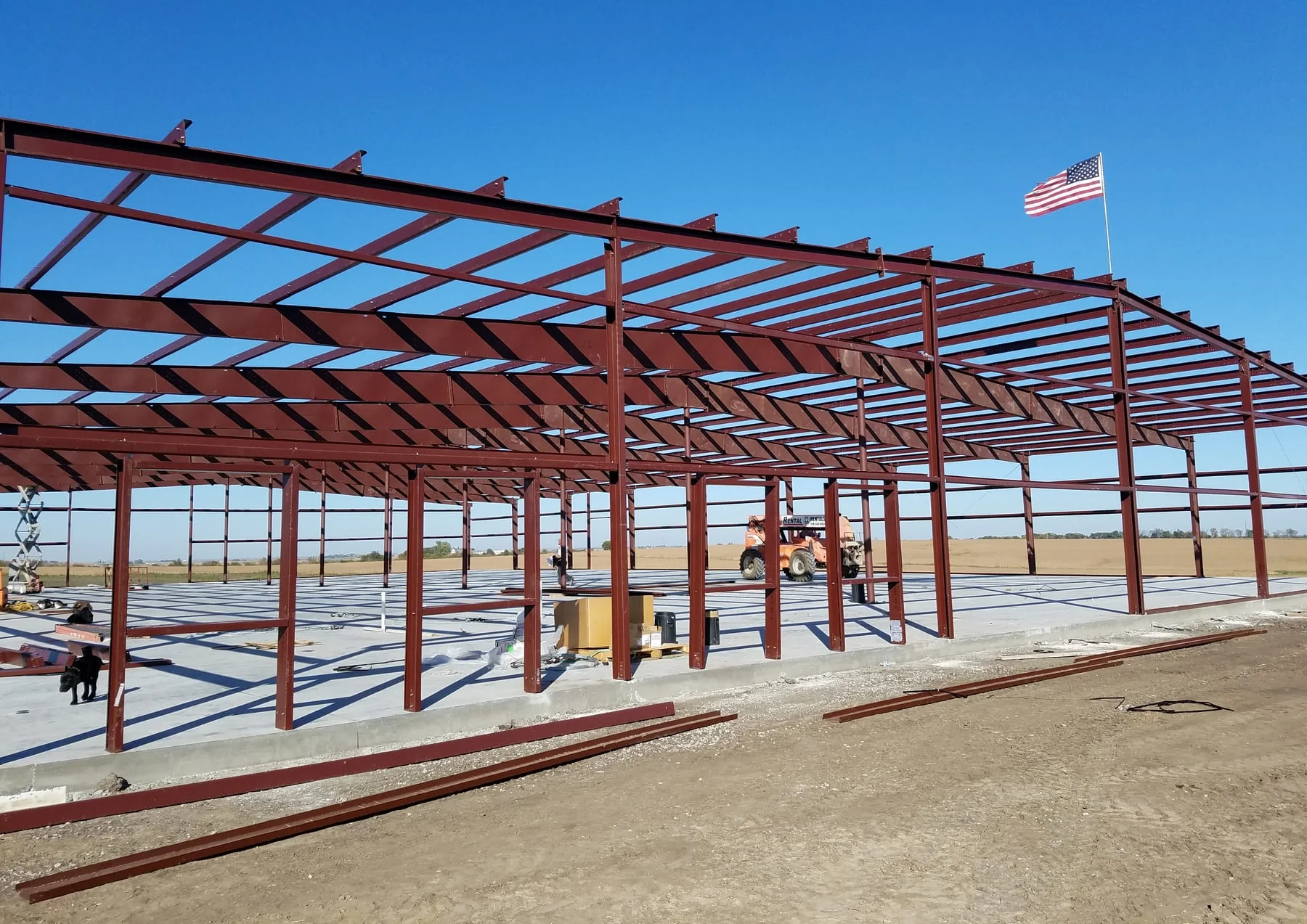 Red steel framing under erection on a concrete slab with an American flag overhead