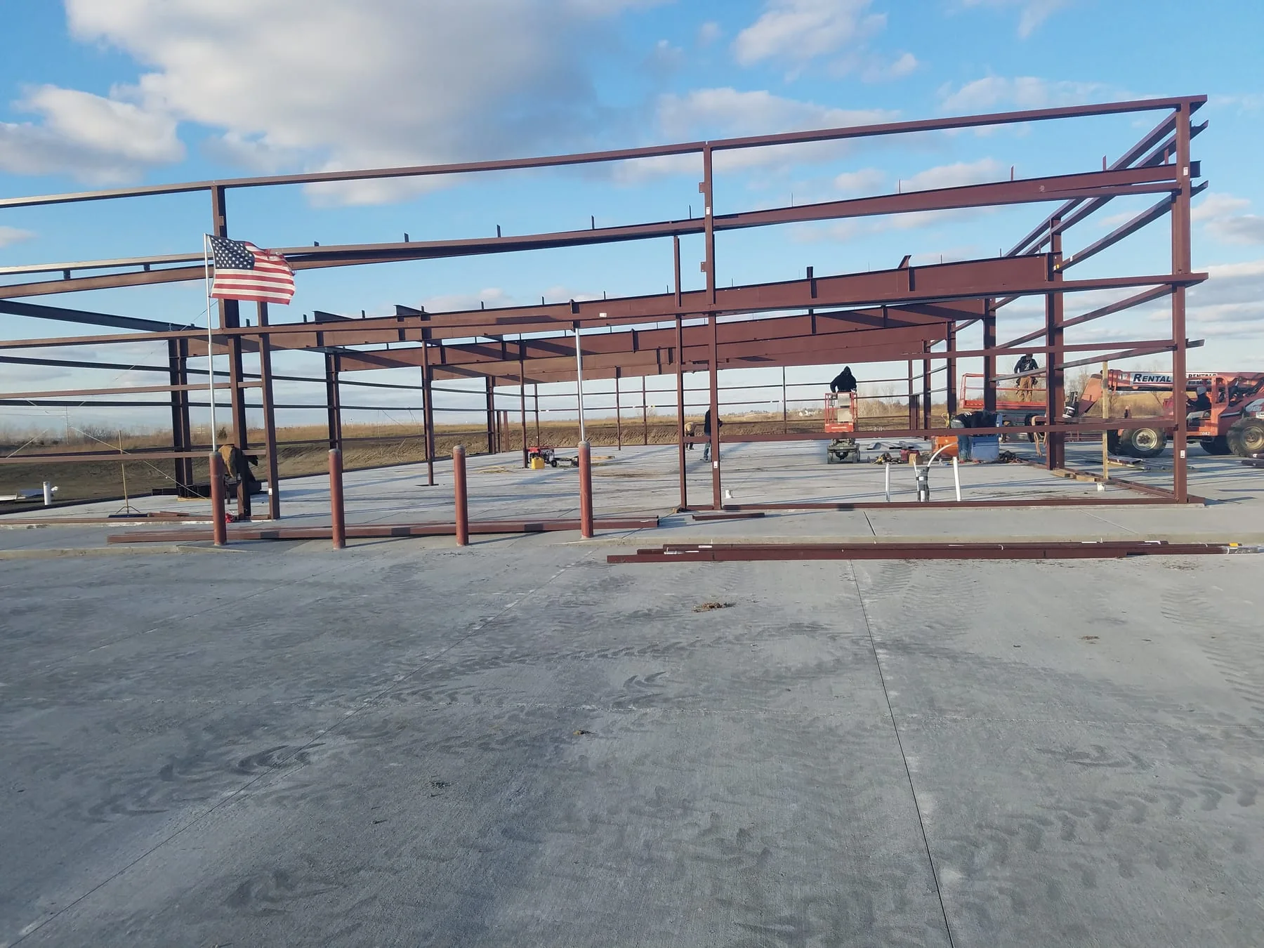 Red steel building frame on a concrete slab with lifts and an American flag