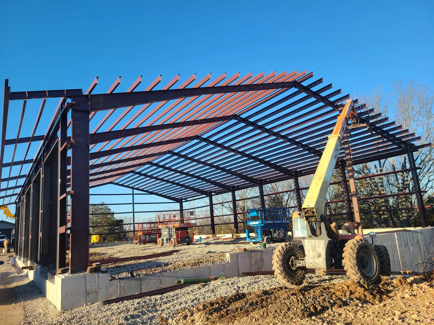 Steel roof framing under construction with lifts and a telehandler nearby