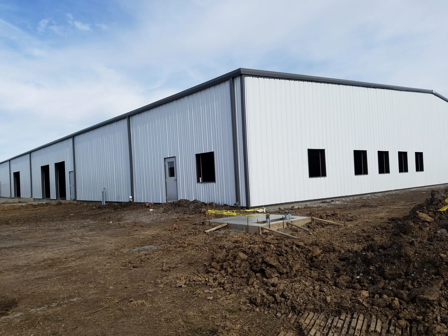 White metal-sided building with dark trim and unfinished ground around the exterior